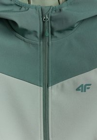 Gros plan d'une veste verte et gris clair avec une fermeture éclair centrale et un logo "4F" sur la partie droite de la poitrine.