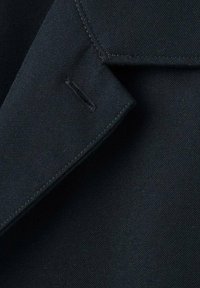 Tela negra con una textura suave, que presenta un cuello puntiagudo y detalles de costura. Incluye un pequeño ojal horizontal.