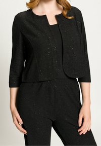 Czarna błyszcząca krótka kurtka z okrągłym dekoltem i rękawami do łokcia, noszona na czarnej bluzce i dopasowanych spodniach. Teksturowany materiał.