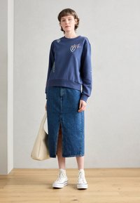 Sweatshirt bleu marine avec des accents brodés ; associé à une jupe en denim avec une fente à l'avant ; baskets montantes blanches ; sac fourre-tout beige.