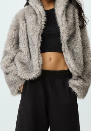 Femme portant une veste en fausse fourrure grise par-dessus un crop top noir et un pantalon noir ample, mains dans les poches, torse visible sur fond blanc.
