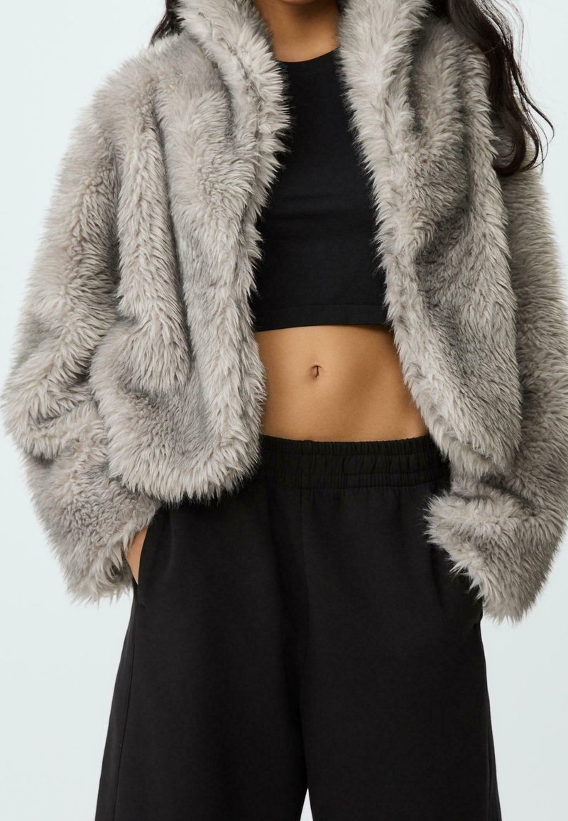 Femme portant une veste en fausse fourrure grise par-dessus un crop top noir et un pantalon noir ample, mains dans les poches, torse visible sur fond blanc.