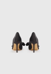 Décolleté in satin nero con punta affusolata e tacchi a spillo sottili. Presentano un dettaglio a fiocco sul retro, con suola beige.