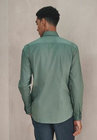 Grünes Langarmhemd aus glattem Stoff, mit klassischem Kragen und geknöpften Manschetten, in einer taillierten Passform sichtbar von hinten.