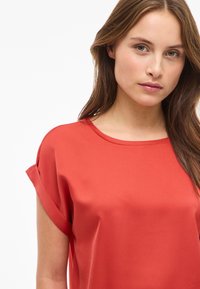 Rød sateng topp med korte ermer og rund halslinning, med rullede mansjetter, som har en glatt tekstur og en løs, avslappet passform.