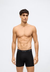 Shirtloze man met kort krullend haar in zwarte Calvin Klein boxer shorts staand tegen een effen lichte achtergrond.