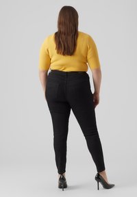 Vaqueros negros de cintura alta con un diseño ajustado. La modelo lleva un top amarillo de canalé y zapatos de tacón negros. Textura de tela suave.