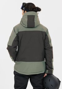 Isolierte Snowboardjacke in Grün und Dunkelgrau, mit Kapuze, kontrastierenden Paneelen und einer glatten, strukturierten Oberfläche. Begleitet von einem schwarzen Helm.