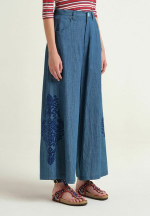Persona che indossa pantaloni blu a gamba larga con ricamo floreale scuro, top a righe rosse e bianche e sandali aperti decorati.