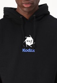 Black hoodie made of soft fabric featuring a white hexagonal logo with "Huf" and "Kodak" in blue on the chest. Drawstring hood included.