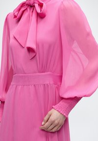 Γυναίκα που φοράει ένα φωτεινό ροζ φόρεμα με μεγάλο φιόγκο στο λαιμό, φουσκωτά διαφανή μακριά μανίκια και ελαστικές μανσέτες και μέση.