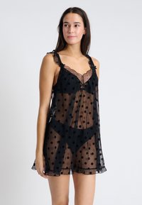 Przezroczysta czarna koszulka nocna w kropki, z falbaniastym dekoltem, marszczonymi ramiączkami i luźnym krojem. Lekka tkanina o przezroczystej strukturze.