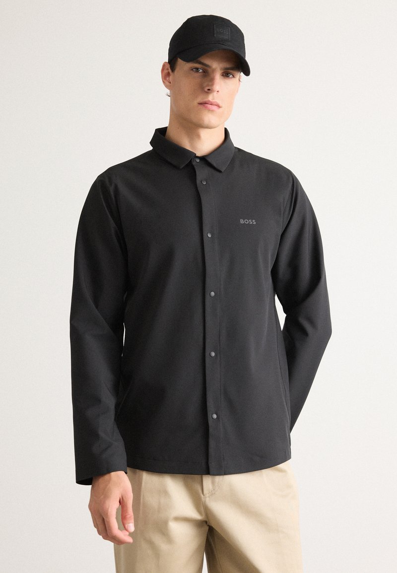 Mann iført svart knappet jakke med BOSS-logo og svart caps, kombinert med beige bukser, står mot hvit bakgrunn.