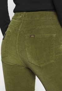 Pantalon en velours côtelé vert olive avec taille haute, présentant des coutures distinctes, deux poches arrière et une étiquette noire avec lettres dorées.