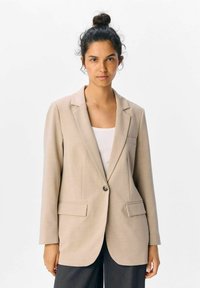 Ljust beige kavaj med slag, enkel knappstängning, två framfickor och ett mjukt, strukturerat tyg. Enkel och modern design.