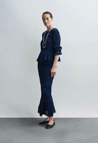 Temno modra peplum majica z teksturiranim tkanjem in napihnjenimi rokavi, združena z oprijeto, razširjeno krilo. Nošena s črnimi ravnimi čevlji in dolgo verižico.