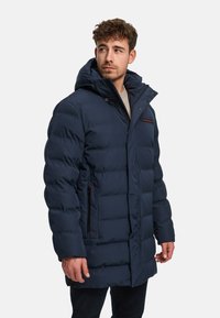 Marineblauwe pufferjas met capuchon, voorzien van een ritssluiting en drukknoopsluiting, geribbelde manchetten en contrasterende rode accenten op de borst en zakken.