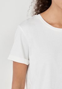 Weißes Baumwoll-T-Shirt mit Rundhalsausschnitt und umgeschlagenen kurzen Ärmeln. Der Stoff wirkt weich mit einer glatten Textur.