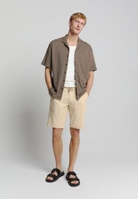 Korte mouwen, bruine shirt met een geometrisch patroon, witte tanktop, beige shorts en zwarte sandalen met gespen.