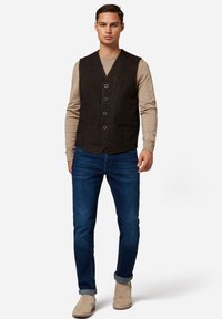 Svart mockaväst med V-ringning, sex knappar, två framfickor, i kombination med en beige långärmad skjorta och blå jeans.