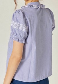 Blouse rayée bleu et blanc avec manches courtes bouffantes, accents floraux brodés sur les épaules et détail de col à volants. Matière en coton.