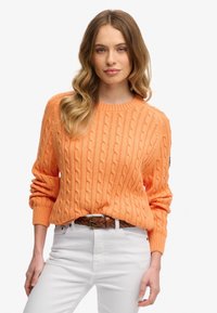 Strikket sweater i lys orange, med rund halsudskæring og ribbede manchetter. Kombineret med hvide bukser og et vævet brunt bælte.