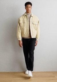 Beige denimjacka med två bröstfickor, beige zip-up tröja, svarta byxor och vita sneakers; står mot en grå vägg.