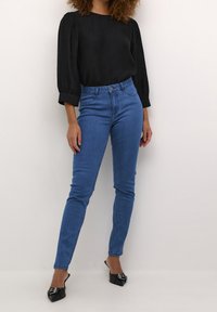 Tumman, tekstuuritetun yläosan, jossa on puffi hihat, paritus korkeavyötäröisten, kapeiden sinisten farkkujen ja teräväkärkisten mustien korkokenkien kanssa.