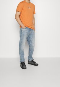 Oranžové bavlněné tričko s bílým logem, modré vybledlé džíny s pohodlným střihem a černé tenisky s texturovanými detaily.