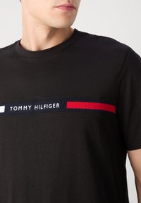Mladić u crnoj Tommy Hilfiger majici s vodoravnom logotip trakom u mornarsko plavoj, bijeloj i crvenoj boji preko prsa.