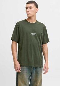 Olivegrünes Baumwoll-T-Shirt mit kurzen Ärmeln, Rundhalsausschnitt und einem kleinen weißen Logo auf der Brust; kombiniert mit hellblauen Jeans.