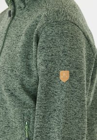 Grön, texturerad fleecjacka med dragkedja fram, en badge på ärmen och en kroppsnära design.