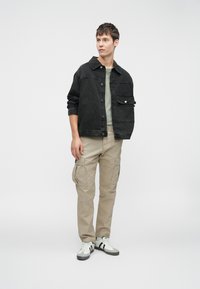 Sort denimjakke med en lomme, olivengrøn t-shirt, beige cargo-bukser og hvide sneakers med sorte striber. Enkel, afslappet outfit.