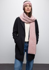 Cappotto di lana nero, maglione crema a trecce e jeans blu. Sciarpa a righe rosa e fascia abbinata aggiungono texture e contrasto di colori.