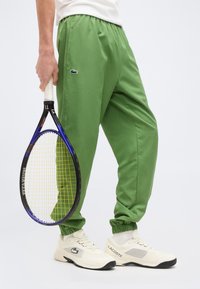 Άτομο που φοράει πράσινο παντελόνι Lacoste και λευκά αθλητικά παπούτσια Lacoste, κρατώντας ένα μοβ και κίτρινο ρακέτα τένις δίπλα του.