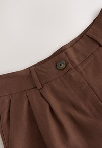 Close-up of brown high-waist pants with a single dark button, belt loop, and pleated fabric detail at the waistband.