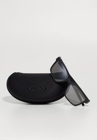 Ochelari de soare negri cu lentile închise, având un design rectangular și un finisaj mat, așezați lângă un toc de protecție negru.