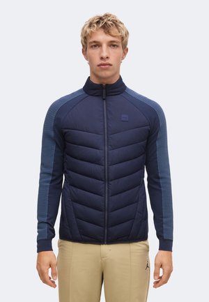 Jeune homme aux cheveux blonds courts et bouclés portant une veste matelassée bleu marine avec des manches rayées et un pantalon beige, debout devant un fond uni.