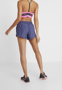 Kvinna sedd bakifrån i rosa Nike sportbh, marinblå träningsshorts och svarta och rosa löparskor mot en vit bakgrund.