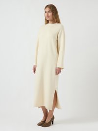 Abito color crema a maniche lunghe realizzato in tessuto morbido, caratterizzato da spacchi laterali e scollo rotondo. Abbinato a scarpe col tacco alte di colore marrone.
