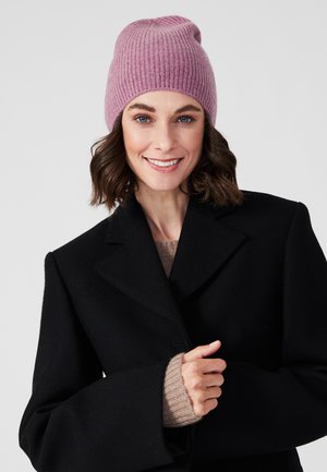 Roze geribde gebreide muts op een model met bruin haar, die een zwarte jas en een beige trui eronder draagt. De muts heeft een losse pasvorm.