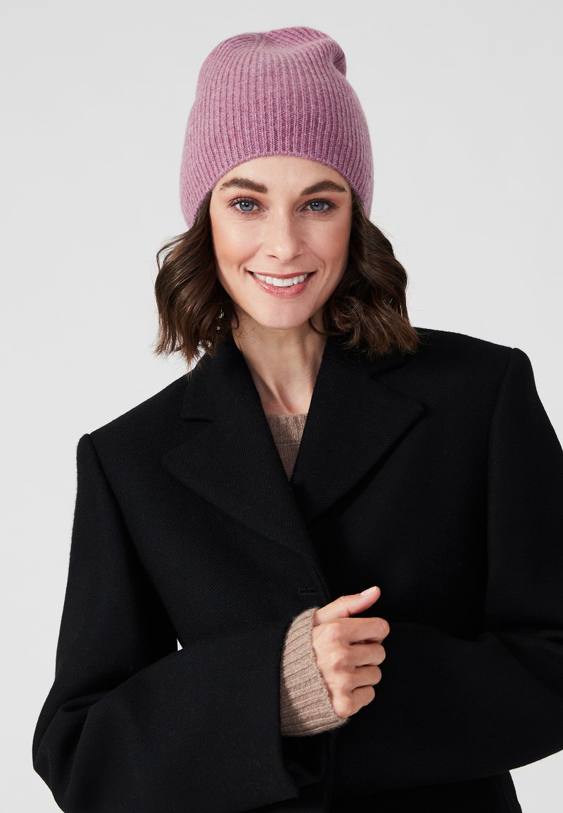 Gorro de punto acanalado rosa en un modelo con cabello castaño, que lleva un abrigo negro y un suéter beige debajo. El gorro tiene un ajuste relajado.