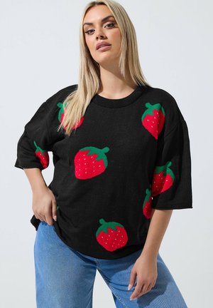 Kvinde med langt blond hår iført en sort sweater med store røde og grønne jordbærmønstre og blå jeans, poserer med hånden på hoften.