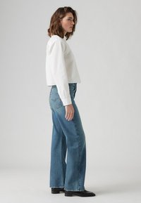 Chemise blanche à manches longues, raccourcie, associée à un jean en denim large bleu clair et des chaussures plates noires. Design simple, tissu en coton texturé.