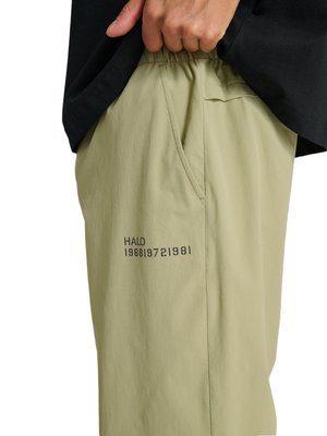 Ręka podciąga jasnobeżowe spodnie z elastycznym stanem z nadrukiem "HALO 196819721981" w pobliżu kieszeni, ubrana w czarną koszulę.