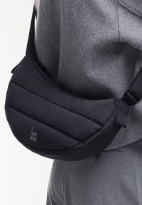 Schwarze, gepolsterte, halbmondförmige Schultertasche mit verstellbarem Riemen, quer über den Oberkörper getragen, über grauer Kleidung.