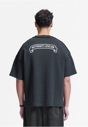 Person med kort krøllet hår iført en løs sort tekstureret skjorte med teksten "RETERNITY ATELIER" på bagsiden, stående mod en hvid baggrund.