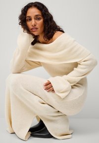 Lange, off-the-shoulder crèmekleurige gebreide jurk met geribbelde textuur en wijde mouwen, gecombineerd met zwarte puntige schoenen.