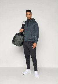 Vīriešu tumši pelēks uzvelkamais kapuce ar kabatām, kombinēts ar melnajām sporta biksēm un baltām sporta apaviem, turēdams melnu trenažieru somu. Vienkāršs, sportisks dizains.