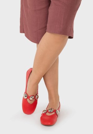 Persona que lleva zapatos planos rojos con grandes decoraciones circulares plateadas, piernas cruzadas y una falda malva hasta la rodilla sobre un fondo blanco.